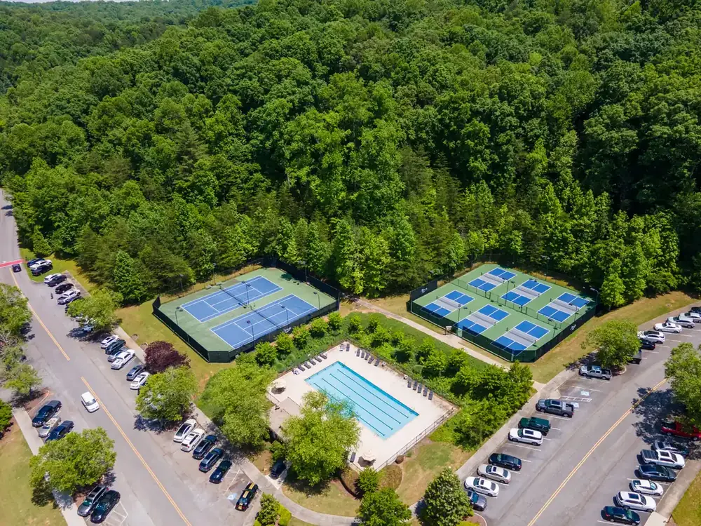 Aerial view of a fenced outdoor pool with lounge chairs beside blue tennis and pickleball courts, surrounded by forest