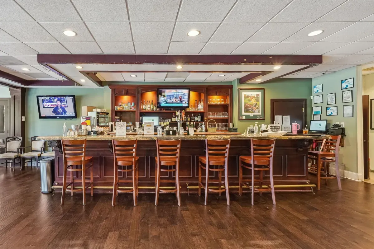Clubhouse bar with a dark wood counter, six wooden bar stools, television screens above the back bar and plank floors