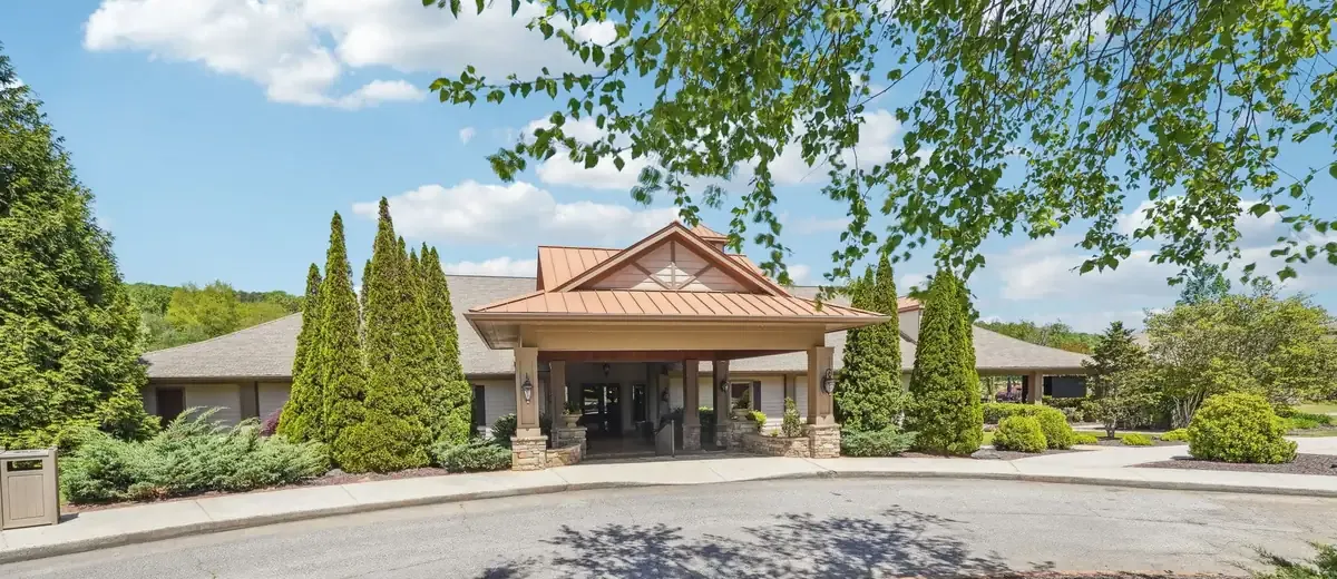Front entrance of the Achasta clubhouse: a copper-roofed portico on stone-based columns between tall evergreens, under a blue sky with leafy branches overhead