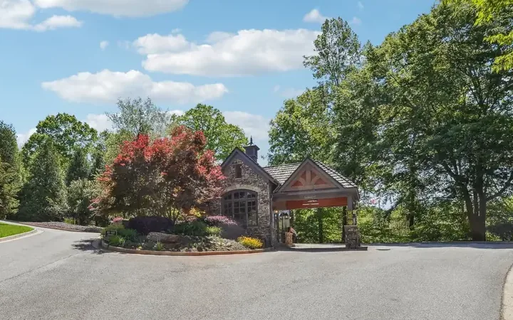 Stone gatehouse with a timber-roofed gate canopy at the community entrance, a red-leaf tree in the landscaped median and a wide paved entry drive