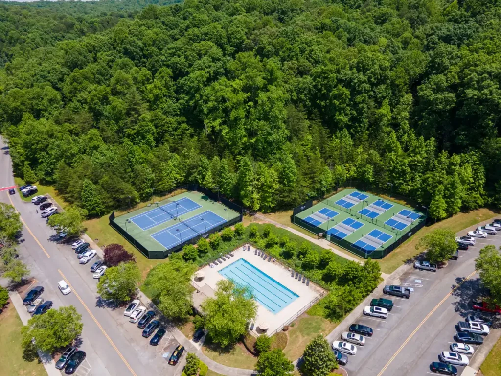 Aerial view of a wooded community amenity area with two blue tennis courts, six blue pickleball courts, and a fenced outdoor lap pool with lounge chairs, surrounded by dense green forest and parking lots lined with cars on a sunny day.