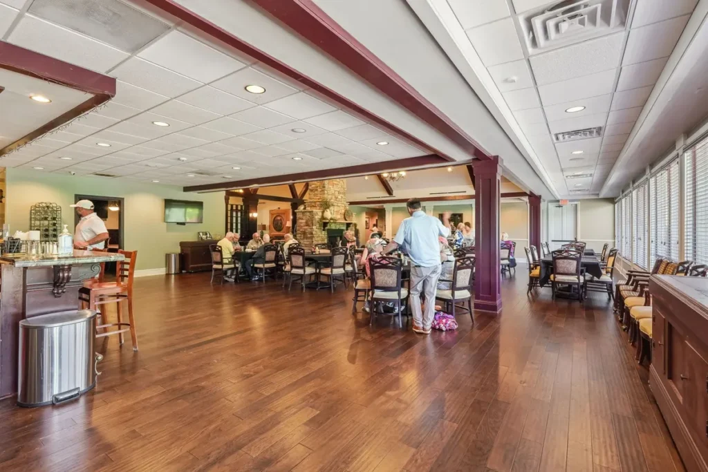 Clubhouse dining hall: hardwood plank floors, columns with wood trim, members seated at dining tables, server station at left, stone fireplace visible in the room beyond.